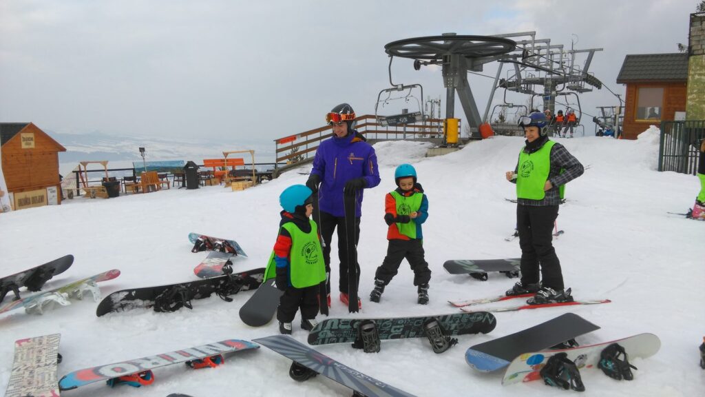 Instruktor z grupą w kamizelkach na śniegu, narty i snowboardy ułożone przed nimi, w tle wyciąg krzesełkowy